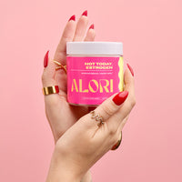 Hand with gold rings holding ALORI Not Today, Estrogen supplement jar against a soft pink background