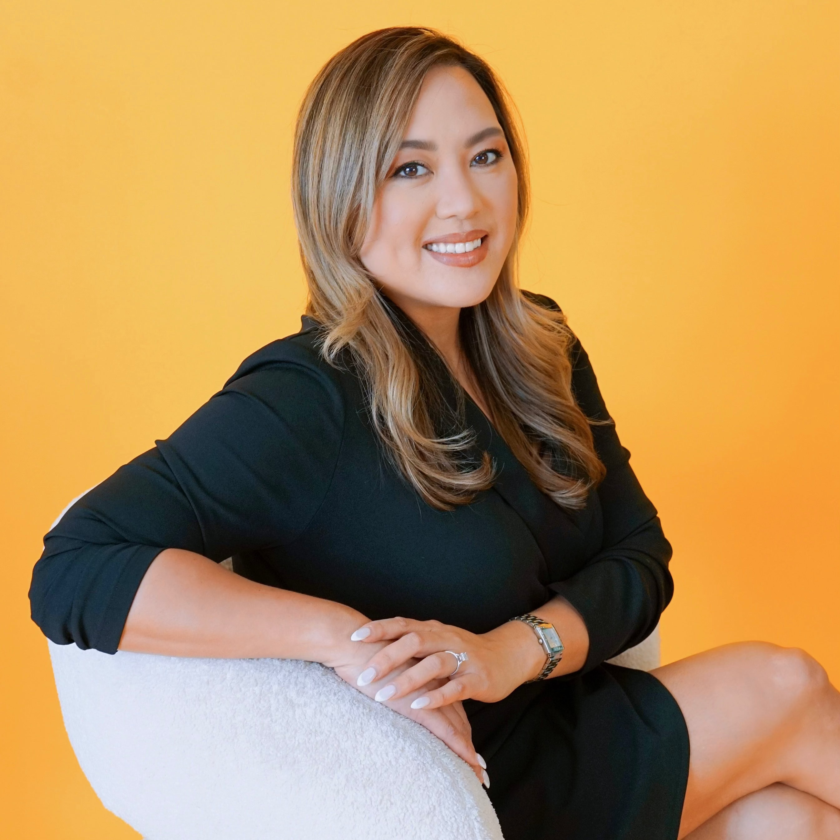 Woman in a black dress sitting on a white chair against an orange background