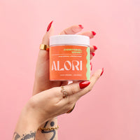 Hand holding ALORI Cortisol, Who? supplement jar against a pink background