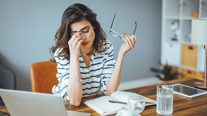 woman sitting at a dStressed woman at her desk rubbing her face and eyes, illustrating the exhaustion, brain fog, and facial puffiness caused by chronically elevated cortisol levelsesk seemingly stressed out working