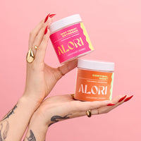 Hands holding ALORI Not Today, Estrogen and Cortisol, Who? supplement jars against a pink background