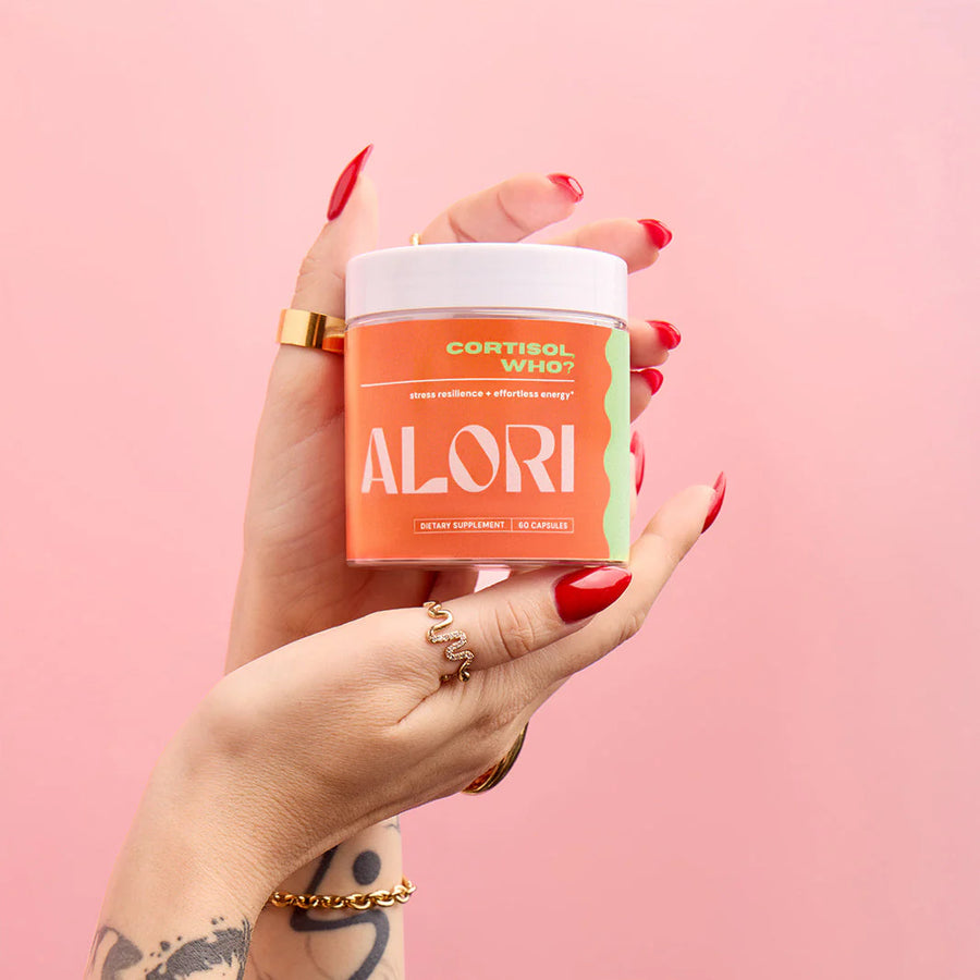 Hand holding ALORI Cortisol, Who? supplement jar against a pink background